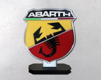 May include: A shield-shaped Abarth emblem sign featuring a black scorpion graphic centered over a yellow and red background. The top section displays the text ABARTH in white letters on a black field, with a thin green, white, and red horizontal stripe below it. The entire emblem is mounted on a black rectangular base.