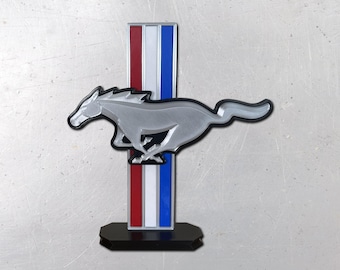 May include: A silver-colored running horse emblem mounted horizontally over a vertical rectangular bar featuring three distinct stripes in red, white, and blue. The entire assembly is supported by a solid black base, creating a display piece.