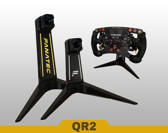 May include: Two black Fanatec display stands with tripod bases and a black carbon fiber racing steering wheel mounted on a third stand. The taller stand features yellow Fanatec text, while the shorter one has a white logo. The text QR2 is visible.