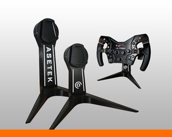 May include: Two black Asetek branded display stands with tripod bases and a black racing steering wheel featuring orange accents and the text Asetek SimSports La Prima. The stands have vertical columns with white stripes and the Asetek logo.