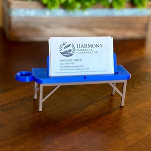 May include: A blue and silver business card holder shaped like a massage table. The holder displays business cards with the text "HARMONY MASSAGE & CHIROPRACTIC" and contact information. The holder is on a wooden surface.