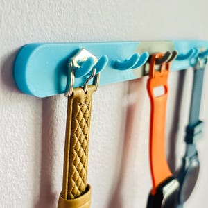 May include: A light blue wall-mounted rack with multiple curved hooks holding three watch bands. The bands include a tan quilted leather strap, a bright orange silicone strap, and a dark blue silicone strap with a watch face attached.