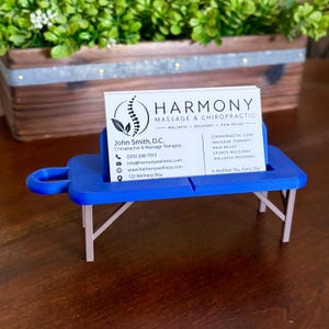 May include: A blue plastic business card holder shaped like a portable massage table with light brown legs. A white business card for Harmony Massage & Chiropractic is inserted into the slot, displaying contact details and service information.