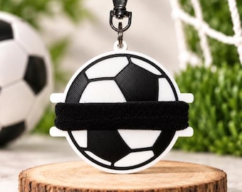 May include: A circular white keychain featuring a black and white soccer ball graphic with a horizontal black elastic hair tie wrapped across the center. The keychain includes a black metal lobster clasp attached to the top for secure fastening.