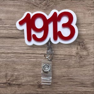 May include: A white and red badge reel with the number "1913" in red. The reel has a clear plastic strap and a metal clip. The background is a wooden surface.