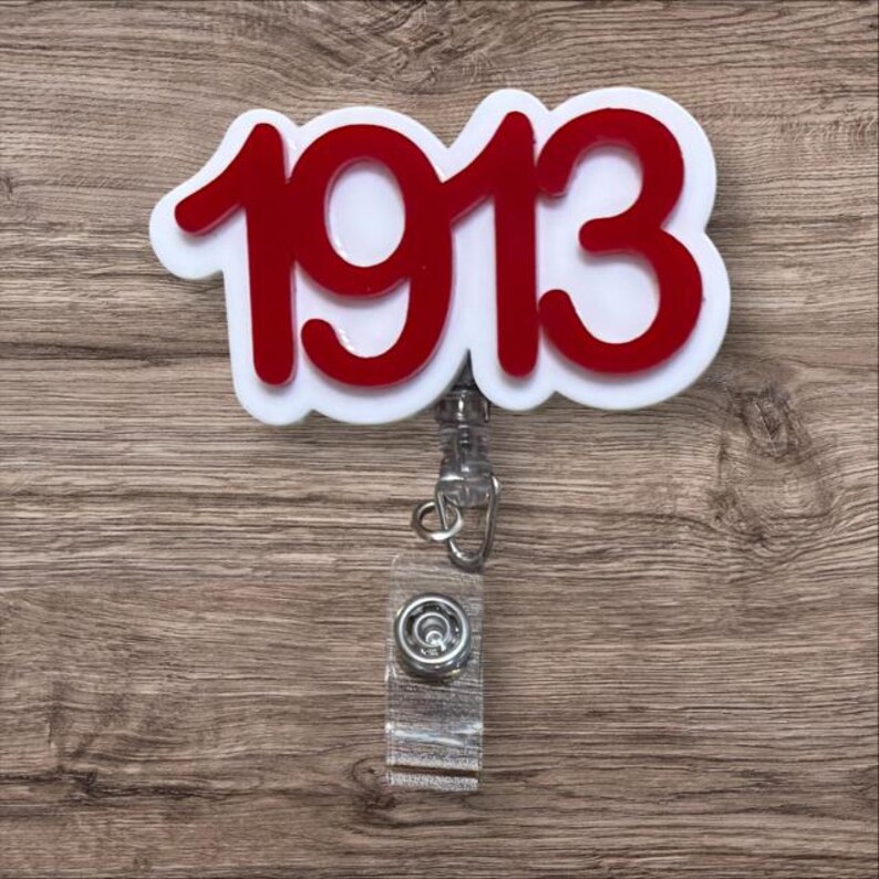 May include: A white and red badge reel with the number "1913" in red. The reel has a clear plastic strap and a metal clip. The background is a wooden surface.