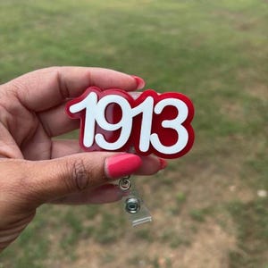 May include: A red and white badge reel with the year "1913" in a bold, stylized font. The badge reel has a clear plastic retractable cord and a metal clip for attachment. The background is a blurred green lawn.
