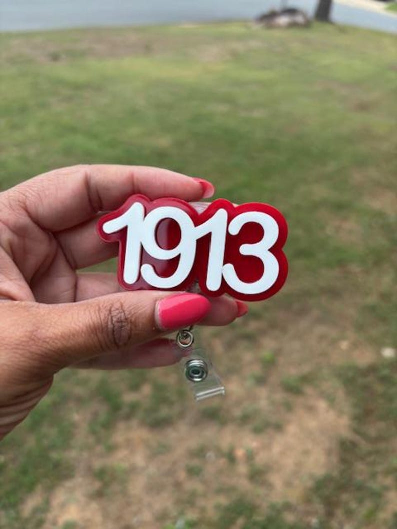 May include: A red and white badge reel with the year "1913" in a bold, stylized font. The badge reel has a clear plastic retractable cord and a metal clip for attachment. The background is a blurred green lawn.