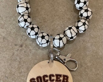 May include: A wooden keychain with a soccer ball design. The keychain has a wooden charm with the words "Soccer Mom" and a soccer ball graphic.