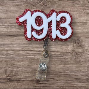May include: A red glitter badge reel with the white number "1913" on it. The badge reel has a clear plastic strap and a metal clip. The background is a wooden surface.