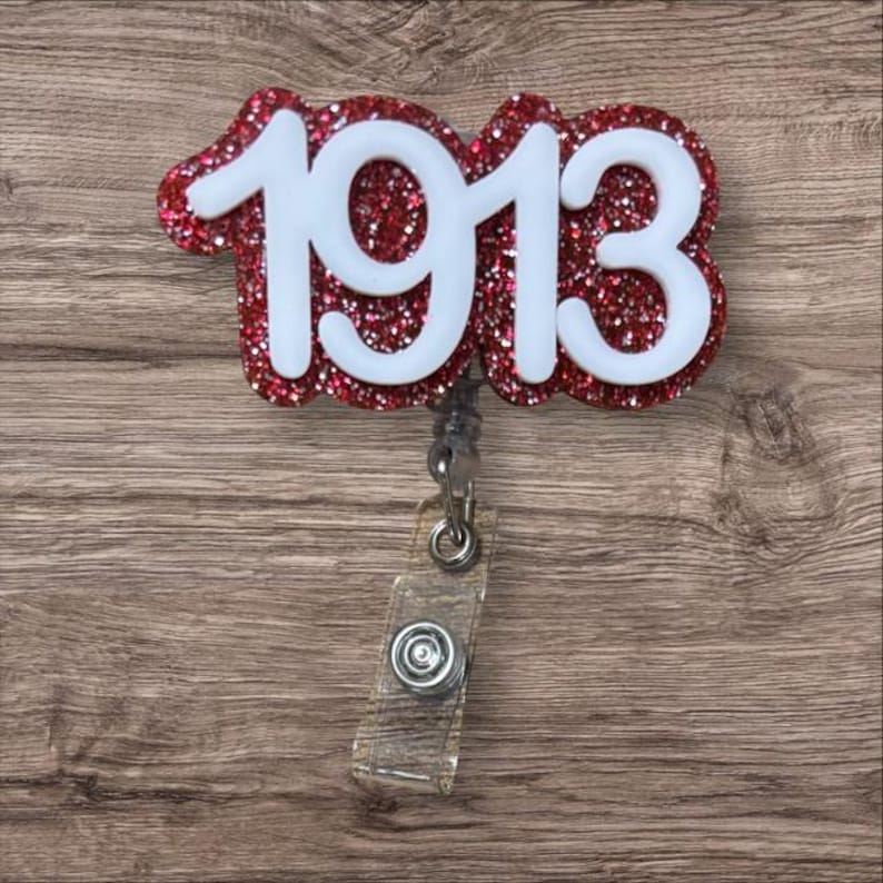 May include: A red glitter badge reel with the white number "1913" on it. The badge reel has a clear plastic strap and a metal clip. The background is a wooden surface.