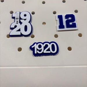 May include: White and blue number and letter charms on a light beige surface. The charms include the numbers 12, 19, 20, and the letters Z, B. The charm with the numbers 19 and 20 also has the letters Z and B. The charm with the numbers 1920 is in a blue outline.