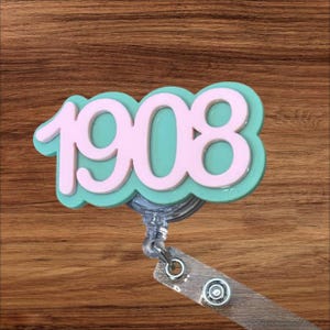 May include: A badge reel with the number "1908" in a layered design. The number is in light pink, outlined in white, and set against a teal background. The reel has a clear plastic body and a wooden-grain clip.
