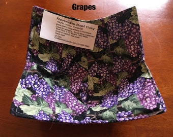 May include: Reversible bowl cosy made from 100% cotton material. The cosy features a black background with a purple grape pattern. The cosy is 10 inches square and is hand-made. It is suitable for small or medium size bowls. Hand wash only. "CraftyplanetCanada 604-345-9006 (NK)" is printed on the tag.