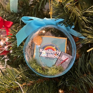 May include: A clear glass Christmas ornament with a blue ribbon and a green moss base. Inside the ornament is a blue and yellow poster with the text "Kimberly Akimbo, a new musical".
