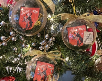 May include: Three clear glass Christmas baubles with gold ribbon and a red and white illustration of a couple dancing with the text "The Music Man" on each bauble. One bauble has a gold trumpet, one has a gold flute, and one has a gold trombone.