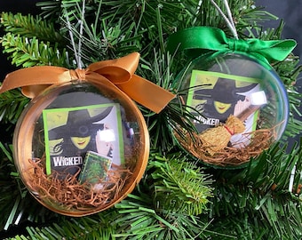 May include: Two clear Christmas ornaments with gold and green ribbons. Each ornament contains a miniature "Wicked" musical poster, a small book, and a miniature broom. The ornaments are hanging on a Christmas tree.