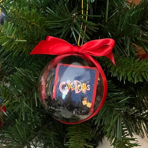 May include: A clear Christmas ornament with a red ribbon bow, hanging on a green Christmas tree. Inside the ornament is a small card with the words "Guys and Dolls" and a cityscape design. The ornament is filled with black decorative material.