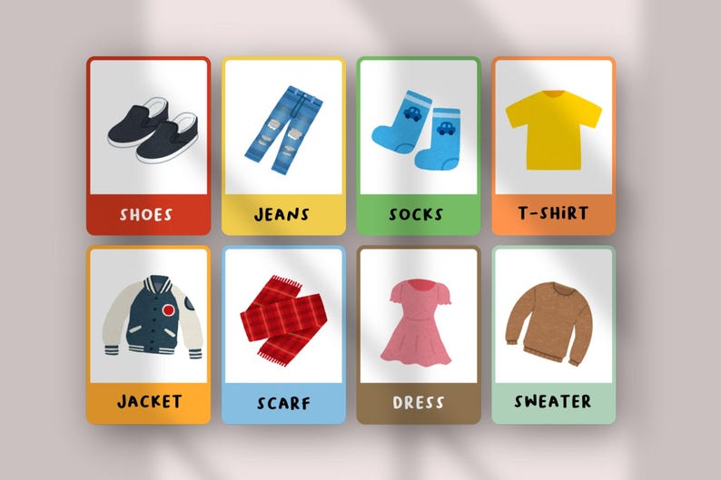 May include: A set of eight educational flashcards featuring illustrations of clothing items including black shoes, blue jeans, blue socks, a yellow t-shirt, a varsity jacket, a red plaid scarf, a pink dress, and a brown sweater with labels.