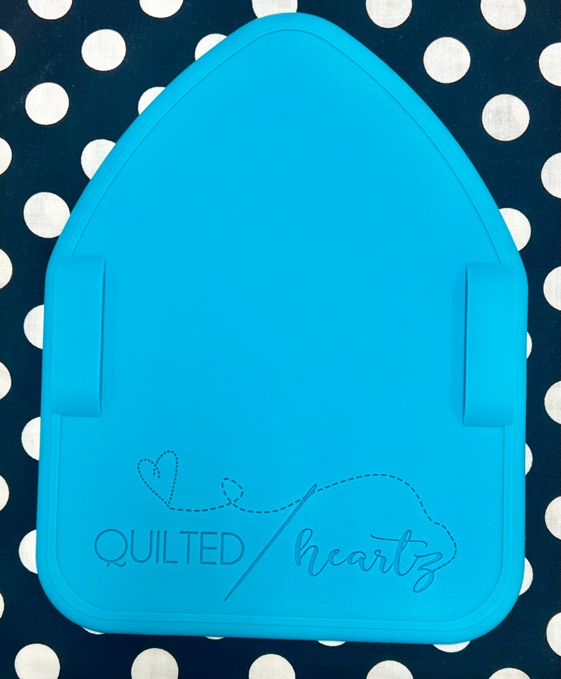 K&ouml;nnte beinhalten: Eine hellblaue Silikon-B&uuml;gelmatte mit einem Herz- und Nadel-Design und dem Text "QUILTED heartz".
