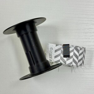 May include: A black plastic spool positioned next to a roll of gray and white chevron patterned ribbon. The ribbon is secured with a small black metal clip and features the printed text "&copy;2023 A" along its white fabric edge.