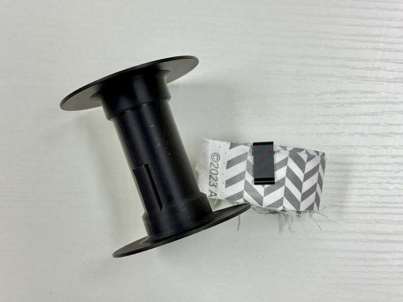 May include: A black plastic spool positioned next to a roll of gray and white chevron patterned ribbon. The ribbon is secured with a small black metal clip and features the printed text "&copy;2023 A" along its white fabric edge.