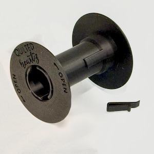 May include: A black plastic bobbin spool featuring the text QUILTED heartz and the word OPEN with an arrow indicator printed on the side. A small, separate black plastic clip is positioned next to the spool on a plain white background.
