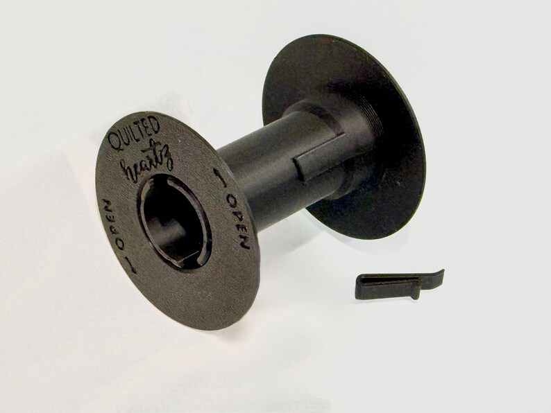 May include: A black plastic bobbin spool featuring the text QUILTED heartz and the word OPEN with an arrow indicator printed on the side. A small, separate black plastic clip is positioned next to the spool on a plain white background.