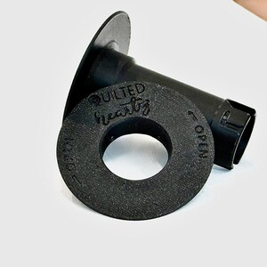 May include: A black 3D-printed spool holder with a textured surface. The circular end cap features the embossed text "QUILTED heartz" and "OPEN" with an arrow indicating the direction to turn for removal from the central cylindrical shaft.