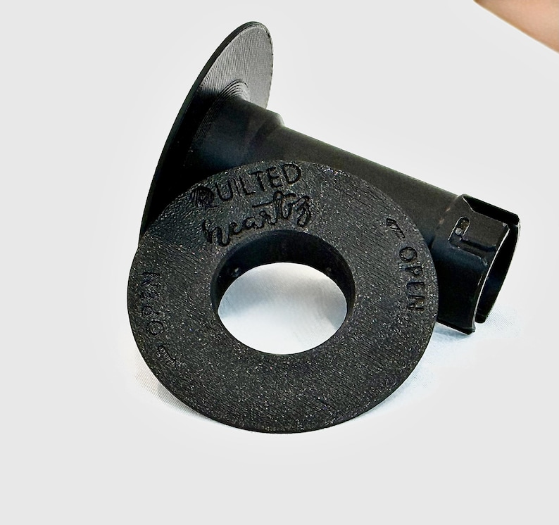May include: A black 3D-printed spool holder with a textured surface. The circular end cap features the embossed text "QUILTED heartz" and "OPEN" with an arrow indicating the direction to turn for removal from the central cylindrical shaft.