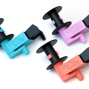 May include: Four spool holders in different colours: turquoise, pink, coral, and black. Each holder has a black spool and the text "QUILTED HEARTZ" printed on the side.