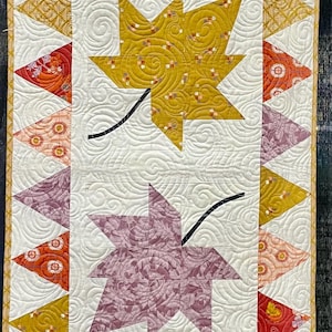 May include: A quilted table runner featuring stylized maple leaves in shades of yellow, orange, and purple. The leaves have black stems and are set against a white background with a swirling pattern. The runner is approximately 15 inches wide.