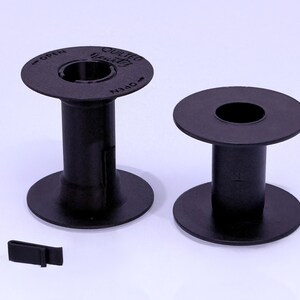 May include: Two black plastic sewing machine bobbins with circular flanges and a central core. The left bobbin features the text "QUILTED" and "OPEN" with directional arrows printed on the top flange. A small black plastic clip is positioned nearby.