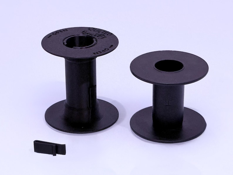 May include: Two black plastic sewing machine bobbins with circular flanges and a central core. The left bobbin features the text "QUILTED" and "OPEN" with directional arrows printed on the top flange. A small black plastic clip is positioned nearby.