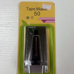 May include: A silver-colored metal bias tape maker tool, labeled Tape Maker 50, encased in clear plastic on a yellow and green cardboard backing. The packaging features a graphic illustration of a folded fabric strip and recycling symbols.
