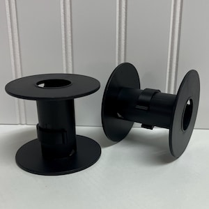 May include: Two black plastic spools with circular end flanges and a central cylindrical core. Each spool features a small rectangular clip attached to the side of the core, designed for securing thread or wire ends during storage or winding.