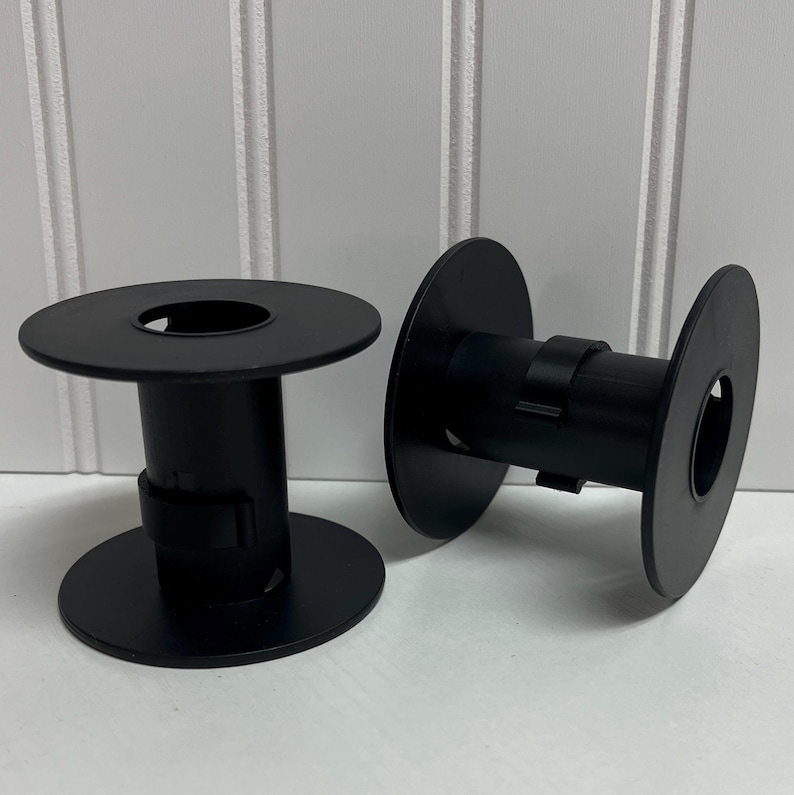 May include: Two black plastic spools with circular end flanges and a central cylindrical core. Each spool features a small rectangular clip attached to the side of the core, designed for securing thread or wire ends during storage or winding.