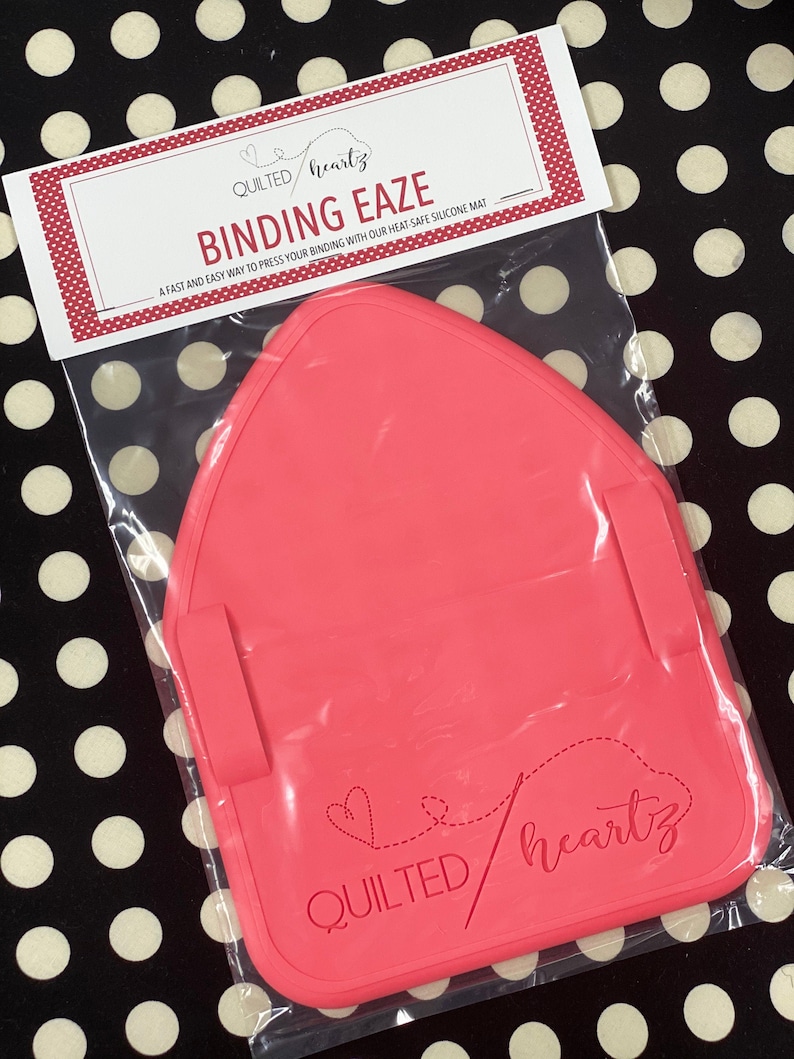 K&ouml;nnte beinhalten: Eine rosa Silikon-Bindungsmatte mit dem Text "QUILTED heartz BINDING EAZE" und einem Herz, durch das eine Nadel geht. Eine schnelle und einfache M&ouml;glichkeit, Ihre Bindung mit unserer hitzebest&auml;ndigen Silikonmatte zu pressen.