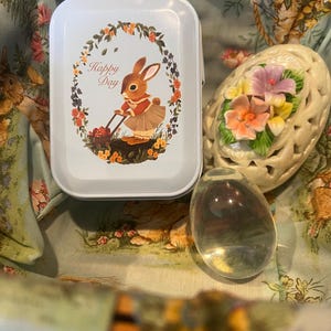 May include: A rectangular light blue metal tin featuring a rabbit illustration and the text Happy Day, a cream-colored ceramic egg with floral accents, and a smooth clear glass egg, all resting inside a fabric-lined basket with a rabbit pattern.