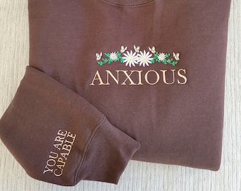 Puede incluir: Sudadera marrón con el texto "ANXIOUS" bordado en dorado en la parte delantera. El texto está rodeado de una corona de flores blancas y hojas verdes. La manga izquierda tiene el texto "YOU ARE CAPABLE" bordado en dorado.