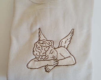 Puede incluir: Una sudadera beige con una imagen bordada en marrón de un ángel durmiendo con alas.