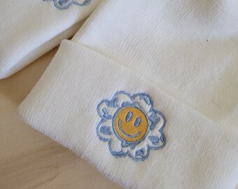 May include: Cream-coloured beanie hat and headband set. Each item has an embroidered design of a yellow smiley face within a blue flower. The beanie has a folded cuff. The items are made of a soft, textured material.
