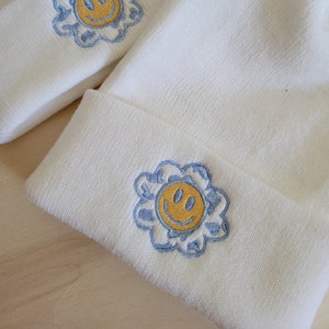 Puede incluir: Conjunto de gorro y diadema de color crema. Cada art&iacute;culo presenta un dise&ntilde;o bordado de una carita sonriente amarilla dentro de una flor azul. El gorro tiene un dobladillo doblado. Los art&iacute;culos est&aacute;n hechos de un material suave y texturizado.