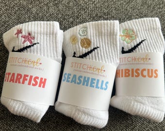 May include: Three pairs of white ankle socks with embroidered designs. One pair features pink starfish, another has seashells, and the third displays a hibiscus flower. Each pair has a black swoosh and a label with the word "STITCHiCal" and the design name.