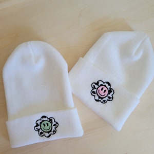 Puede incluir: Dos gorros blancos con dobladillo. Cada gorro presenta un dise&ntilde;o de flor con una carita sonriente en el centro. Una flor tiene una cara verde y la otra una cara rosa, ambas con contornos negros.