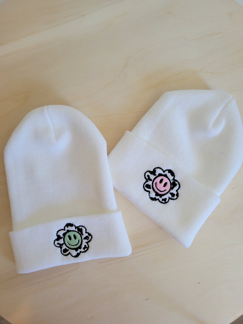 Puede incluir: Dos gorros blancos con dobladillo. Cada gorro presenta un dise&ntilde;o de flor con una carita sonriente en el centro. Una flor tiene una cara verde y la otra una cara rosa, ambas con contornos negros.