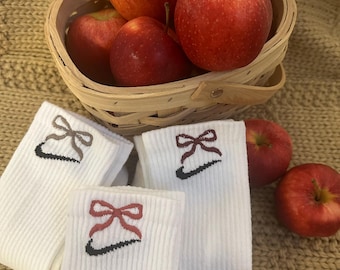 Peut inclure: Un panier en osier contient des pommes rouges. Des chaussettes blanches avec des nœuds brodés et un logo noir sont présentées à côté. L'ensemble est disposé sur une surface beige texturée.
