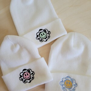 Puede incluir: Tres gorros de punto blancos con dise&ntilde;os de flores bordadas. Cada gorro presenta una cara sonriente de diferente color dentro de la flor: verde, rosa y azul. Los gorros est&aacute;n doblados en el borde.