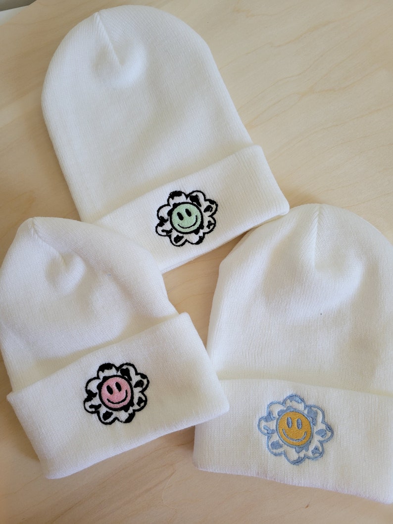 Puede incluir: Tres gorros de punto blancos con dise&ntilde;os de flores bordadas. Cada gorro presenta una cara sonriente de diferente color dentro de la flor: verde, rosa y azul. Los gorros est&aacute;n doblados en el borde.