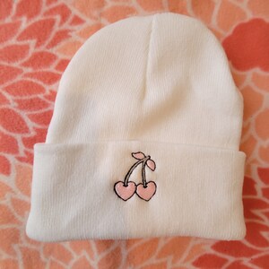 May include: White beanie with a pink cherry embroidered on the front.